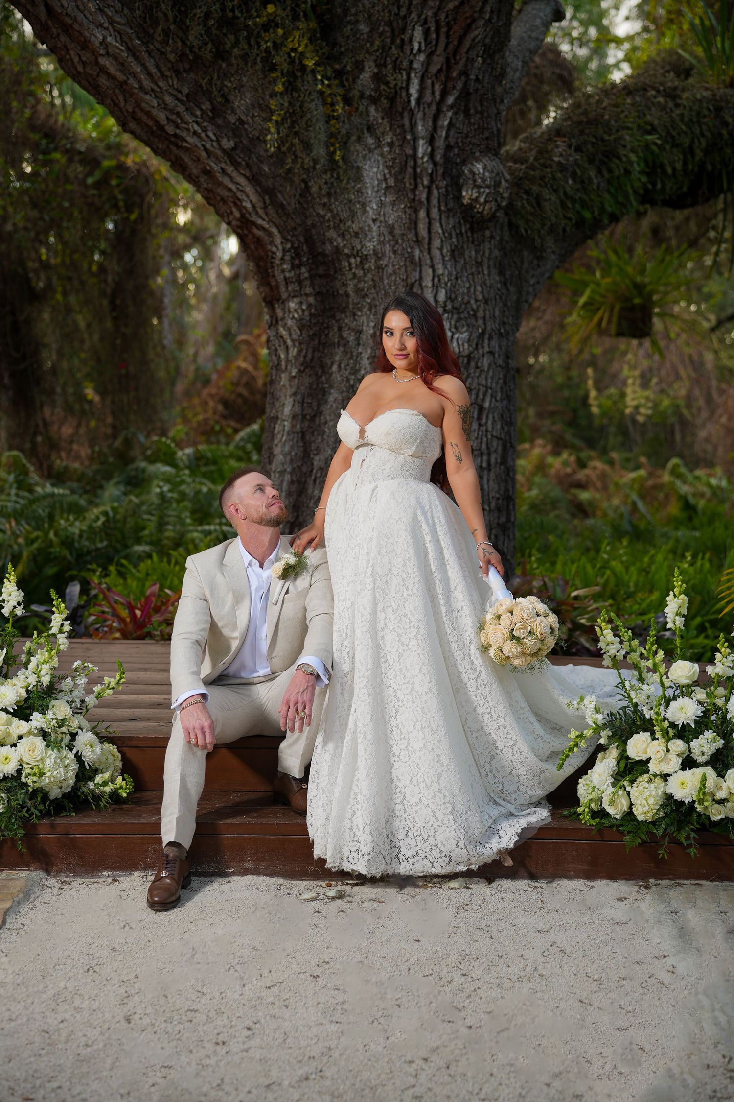 Bride in lace gown holding bouquet stands beside seated groom in tan suit under large tree with white floral arrangements.