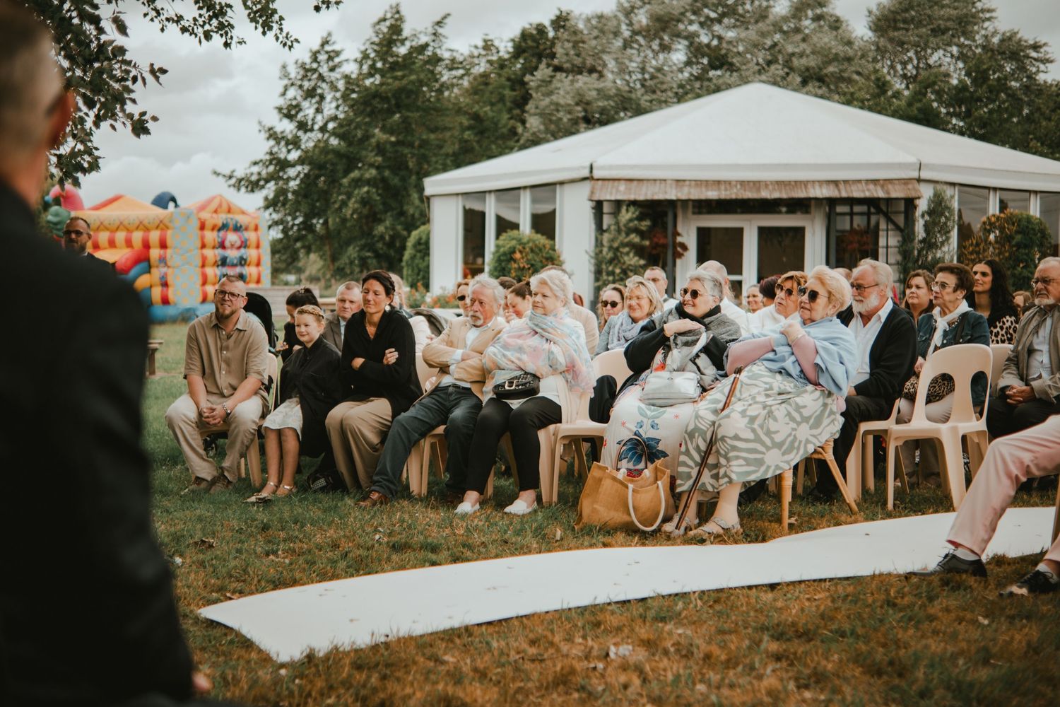 Guests seated outdoors at a wedding ceremony on a lawn with a white tent and trees in the background.