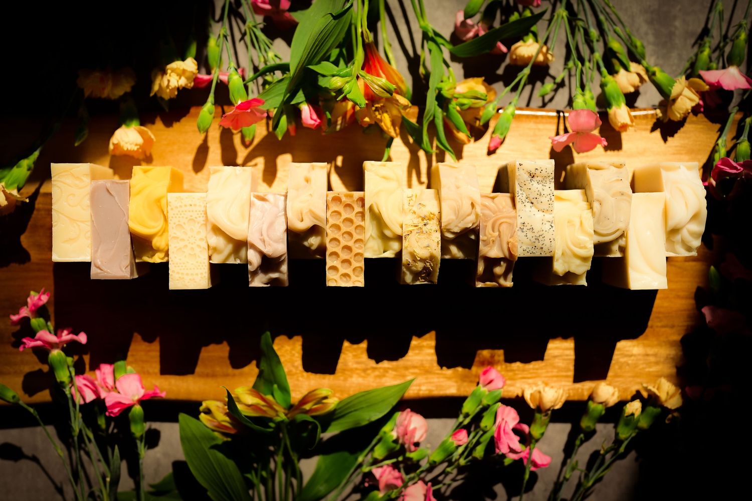 Assorted artisan cheese slices displayed on a wooden board surrounded by colorful pink and yellow flowers.