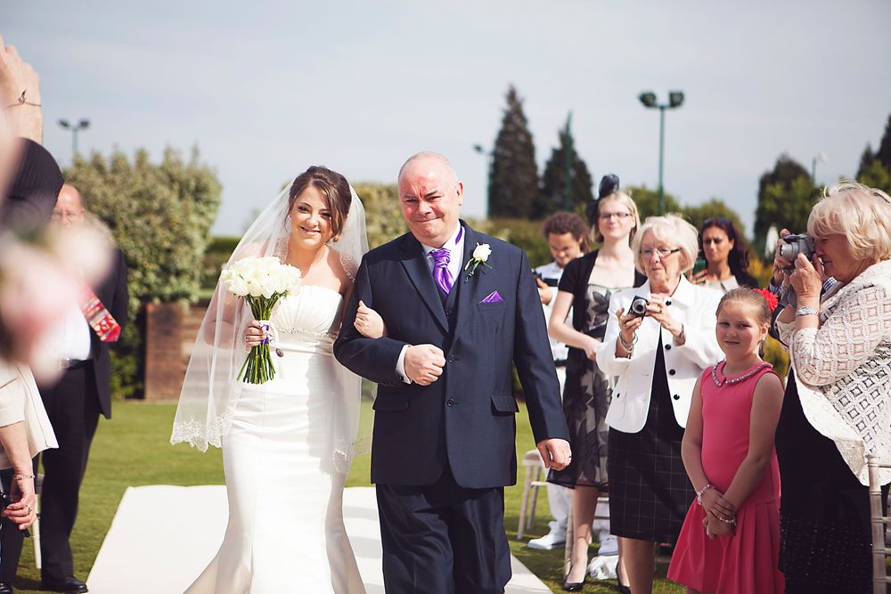 Bride in white gown walks down outdoor aisle with escort at sunny wedding ceremony, guests watching and smiling.