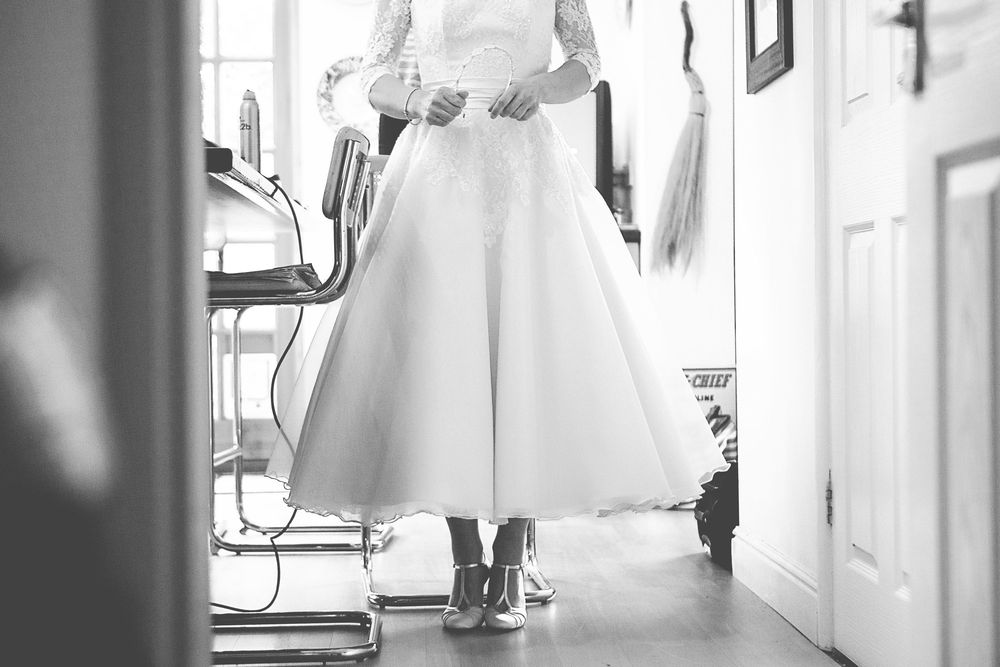 Black and white photo of a bride in a tea-length wedding dress, adjusting her gown in a bright room.