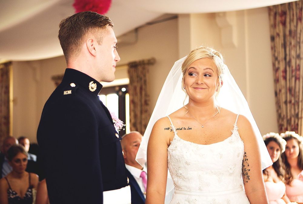 A bride in a white gown and veil smiles at a groom in military dress uniform during a wedding ceremony.