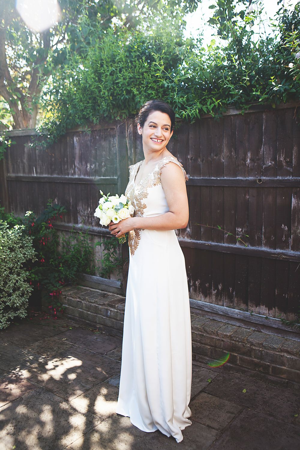 Smiling bride in white gown with gold lace bodice holding bouquet, standing in sunlit garden by wooden fence.