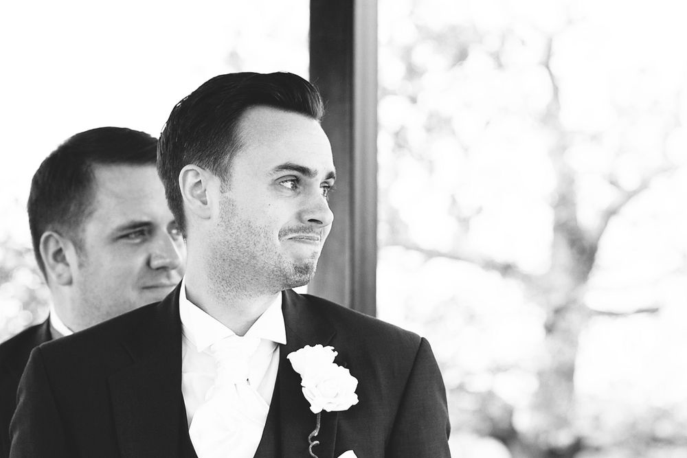 Black and white photo of a groom in a suit with boutonniere smiling at his wedding ceremony.