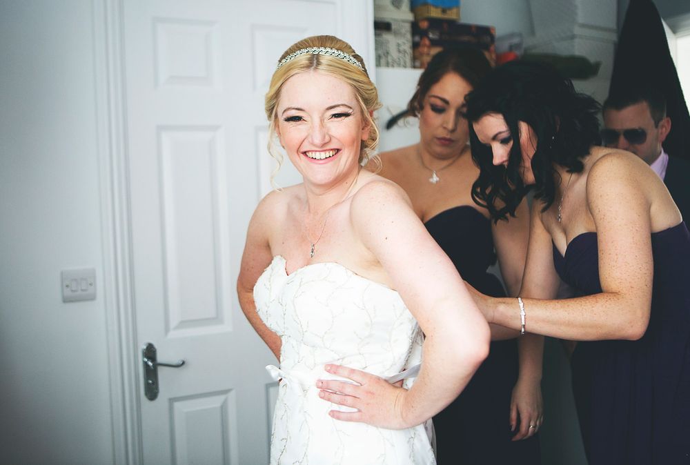 Bride in white strapless gown smiling as bridesmaids in navy dresses help fasten her dress before wedding ceremony.