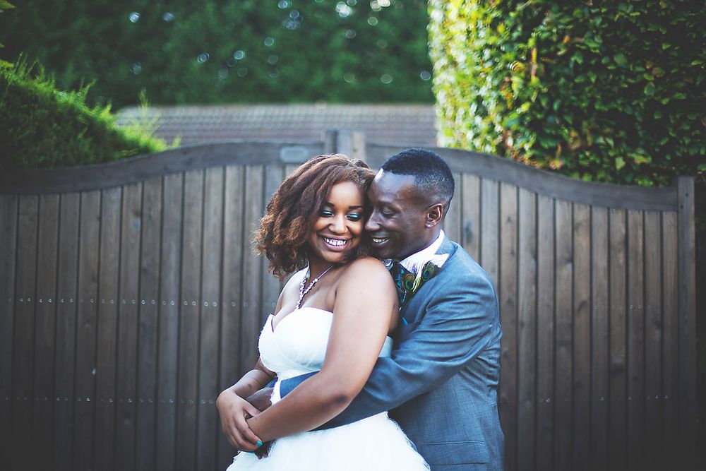 Happy newlywed couple embracing and smiling in front of wooden gates surrounded by lush greenery on their wedding day.