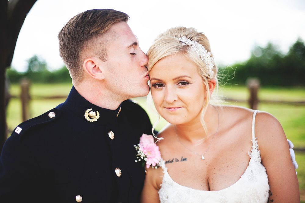 A groom in military uniform kisses the cheek of a smiling blonde bride wearing a lace dress and floral hair accessory.