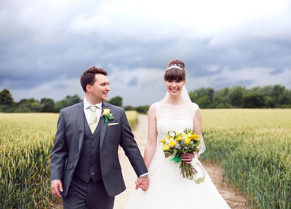 Bride and groom holding hands walking through green fields on their wedding day, bride holding yellow bouquet.