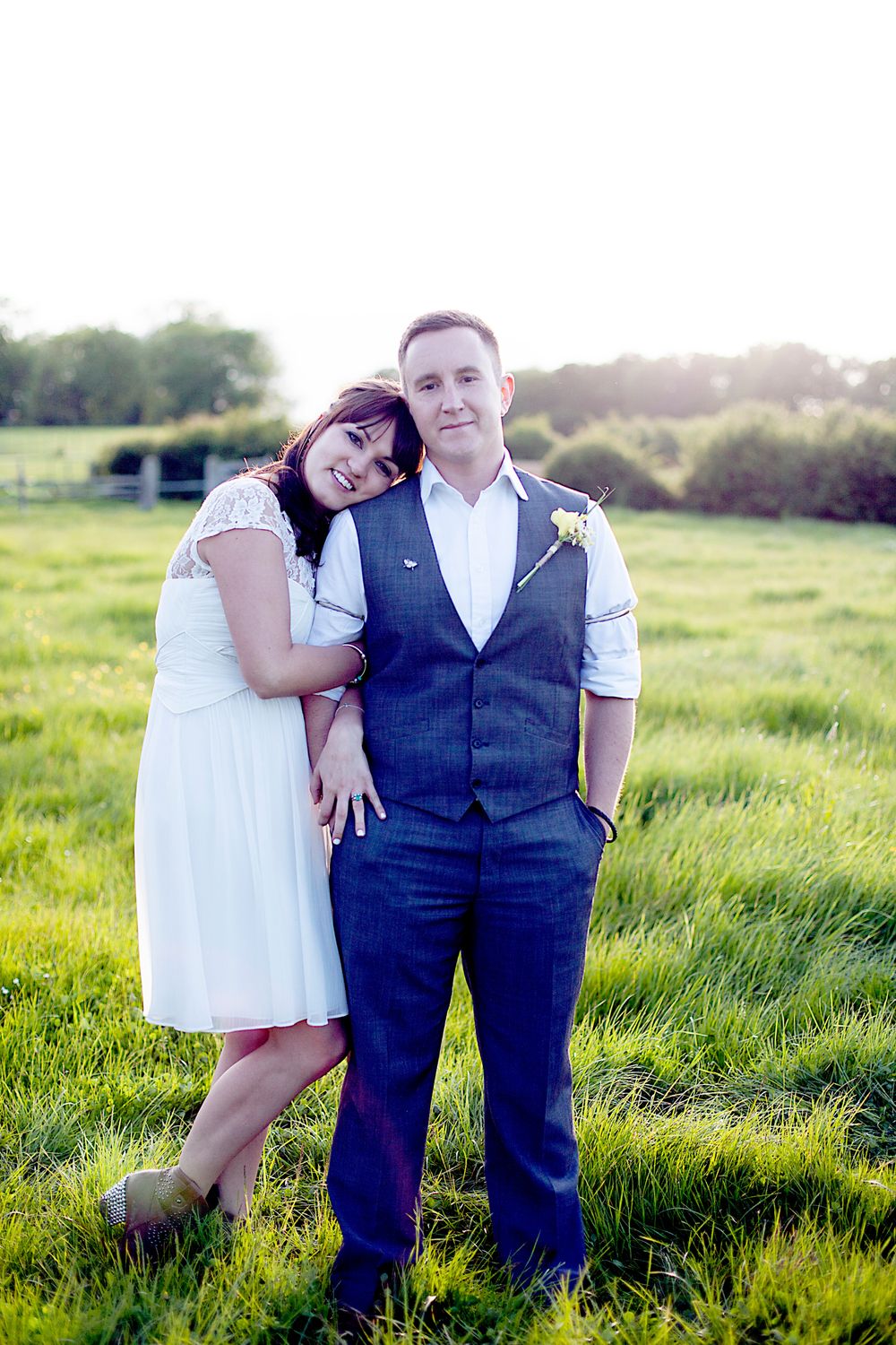 A couple poses in a green field at golden hour, the bride in white dress leaning on the groom in a grey vest suit.
