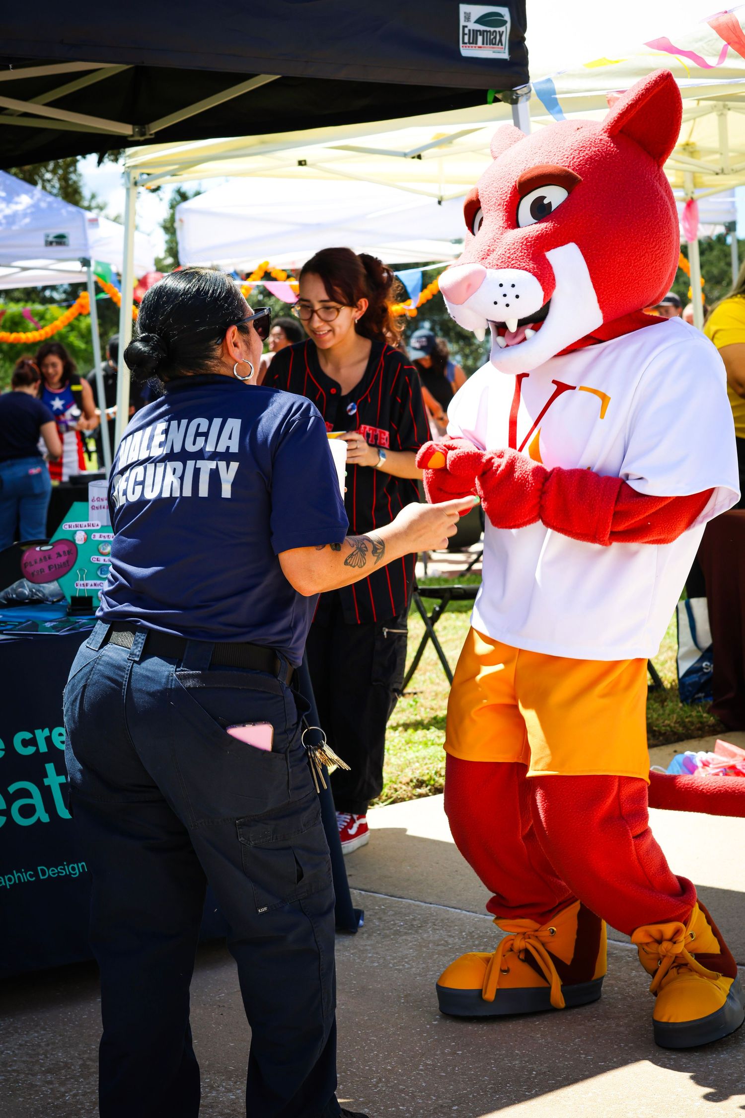 Valencia College mascot in orange costume interacts with security staff at an outdoor campus event booth.