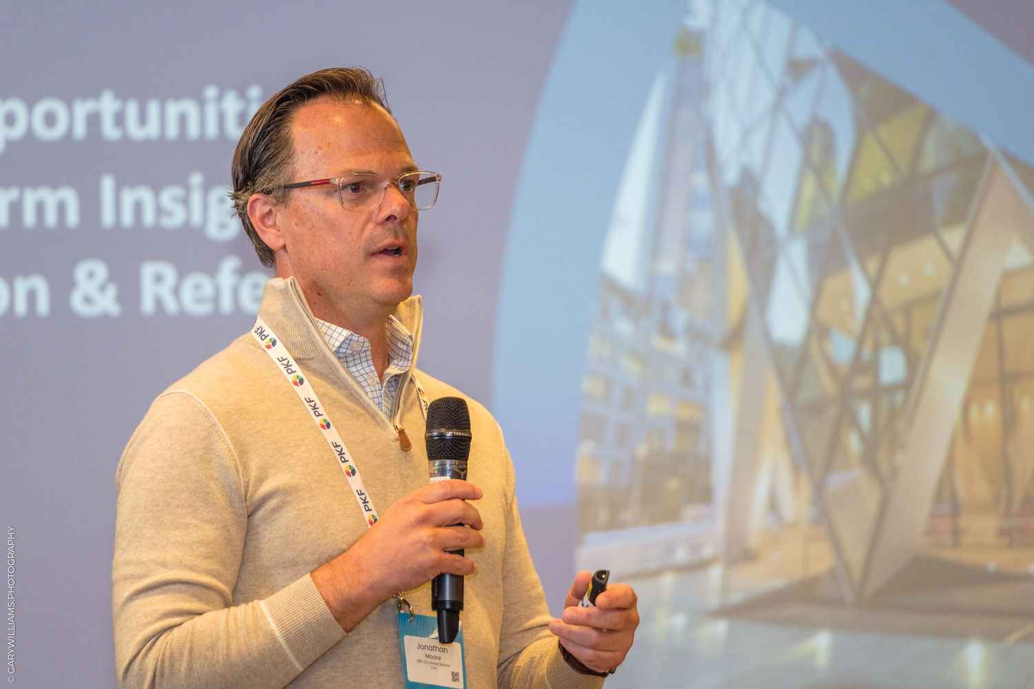 Professional male speaker presenting at a conference, holding a microphone, wearing a beige sweater with a lanyard badge.
