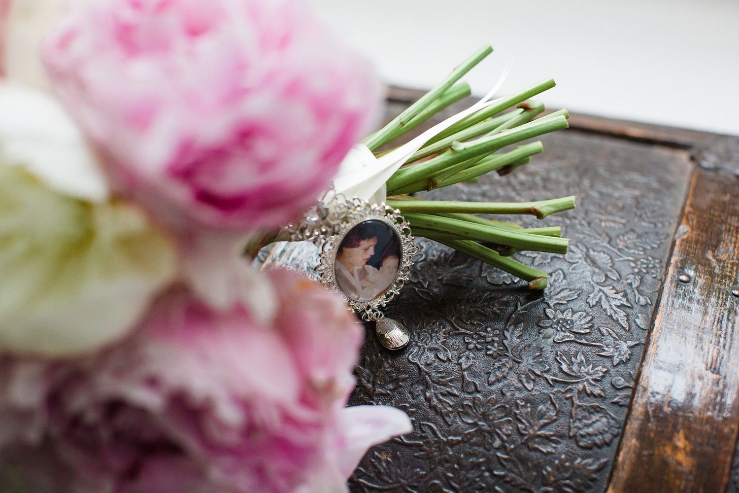 Silver locket charm bracelet beside pink peonies and white floral boutonniere on vintage dark wooden box.