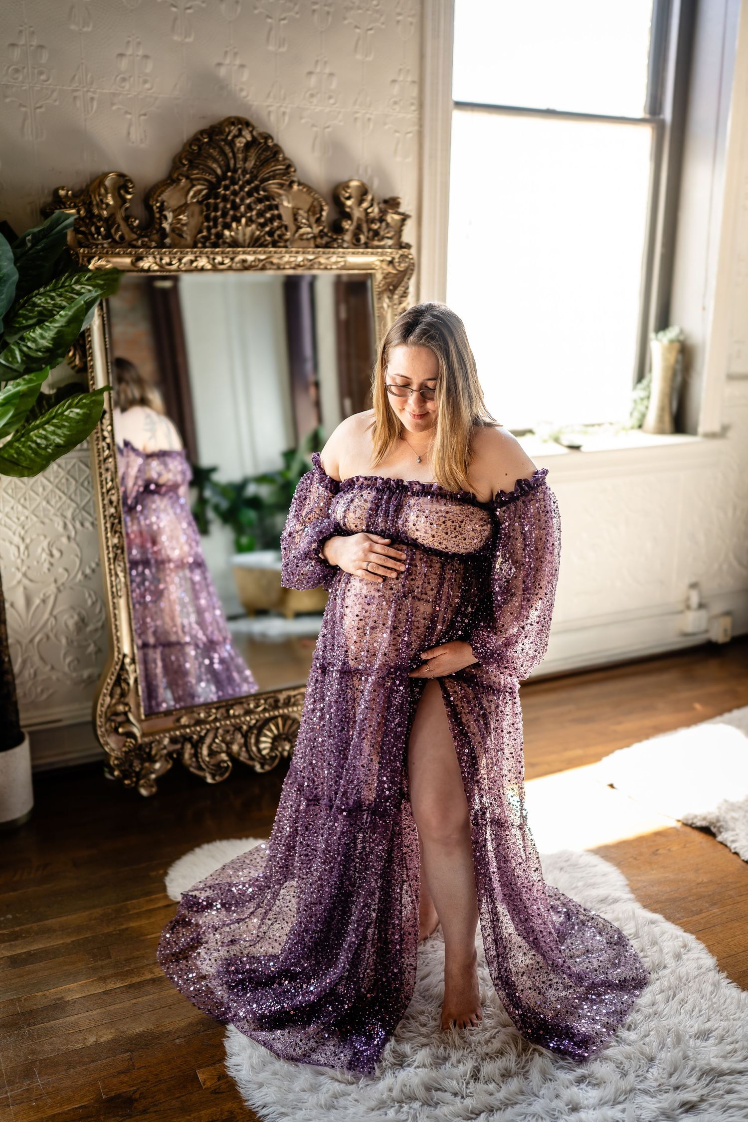 Pregnant woman in purple sequin maternity gown poses in front of gold ornate mirror in bright studio.