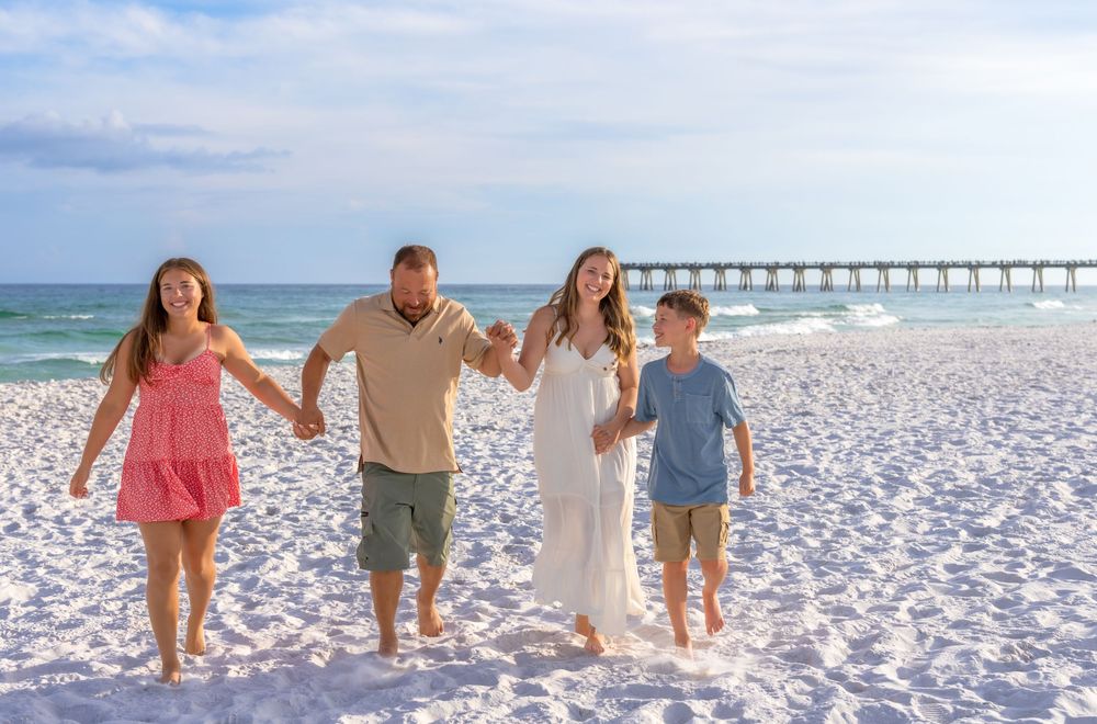 Family of four holding hands walking on a white sand beach with a pier in the background on a sunny day.