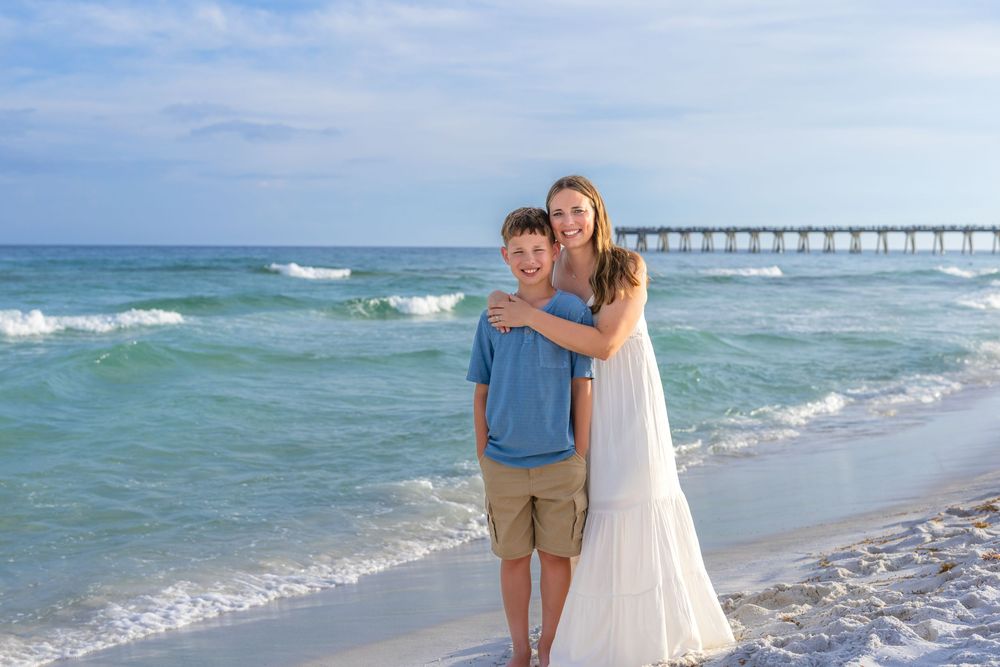 A woman and boy embrace on a sunny beach with turquoise waves and a pier in the background.