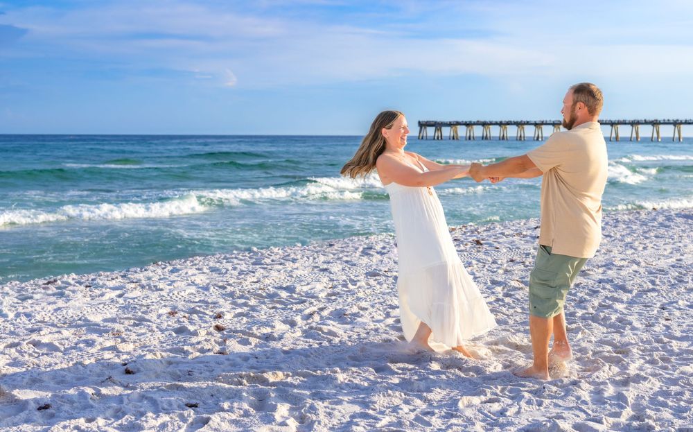 Couple dancing joyfully on a white sandy beach near a pier with turquoise ocean waves in the background.