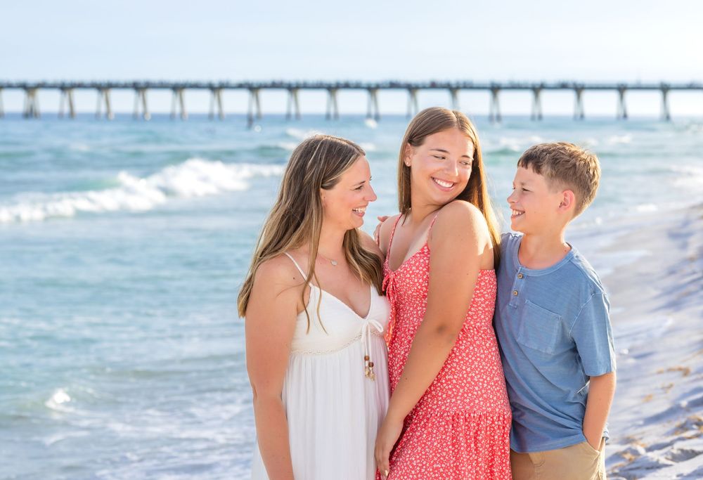 Three smiling teens laughing together on a sunny beach with a pier, two girls and a boy in casual summer outfits.