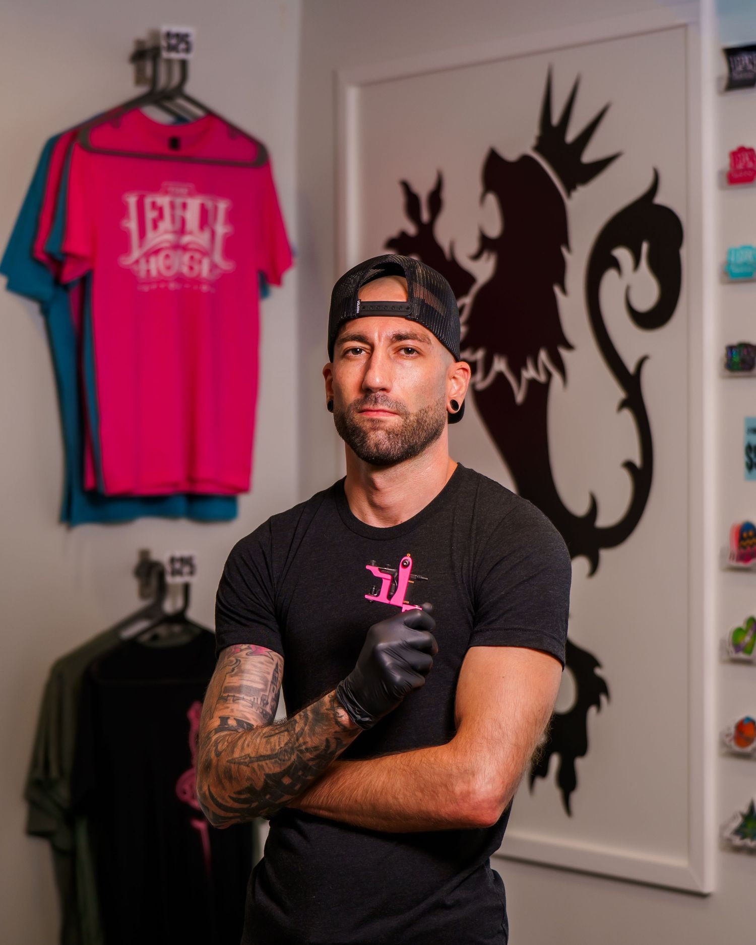 Tattooed man in black cap and gloves poses arms crossed in a merchandise store with pink graphic tees.