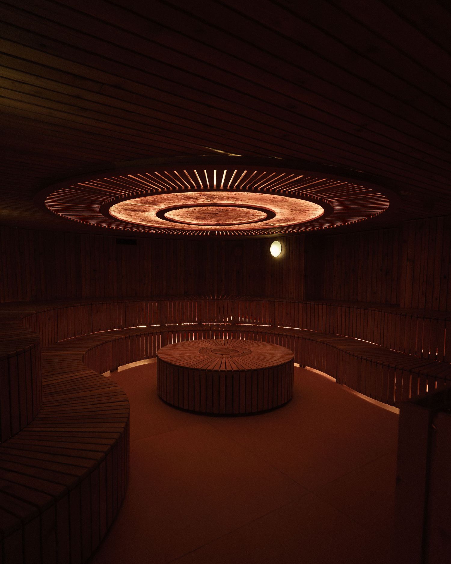Circular wooden sauna with warm amber lighting, tiered benches, and a stunning illuminated ceiling centerpiece.