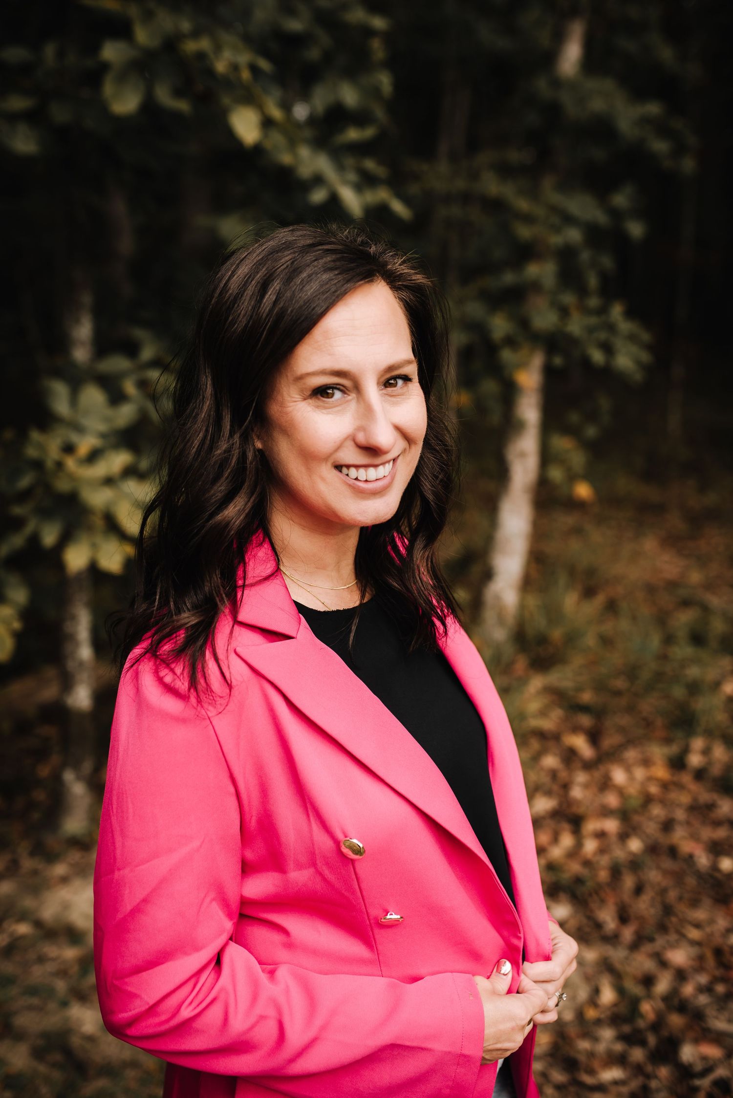 Smiling woman in pink blazer and black top standing outdoors in a wooded setting with autumn leaves.