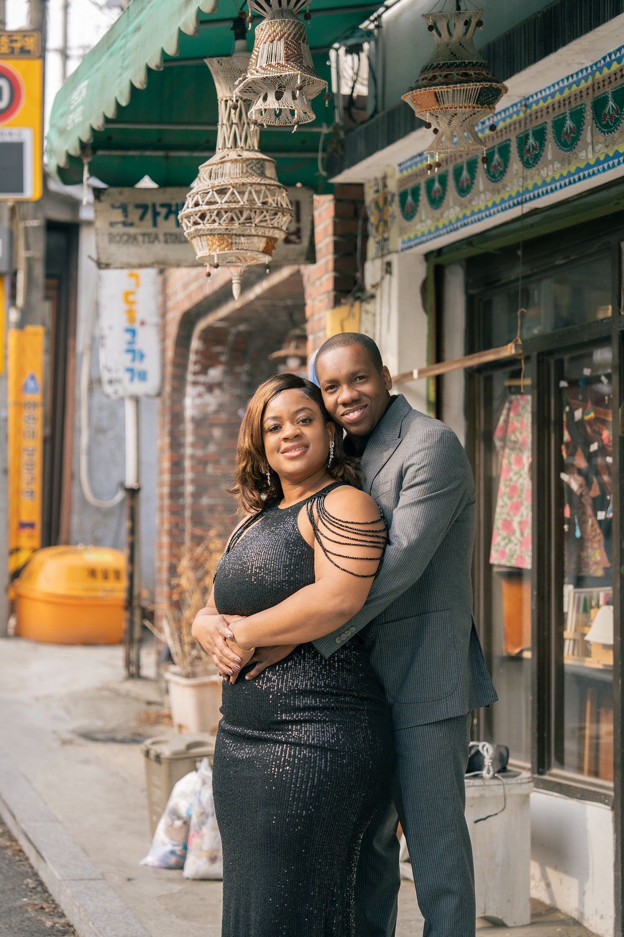 Elegant couple in formal attire embracing on a city sidewalk outside a shop, woman in black gown, man in gray suit.