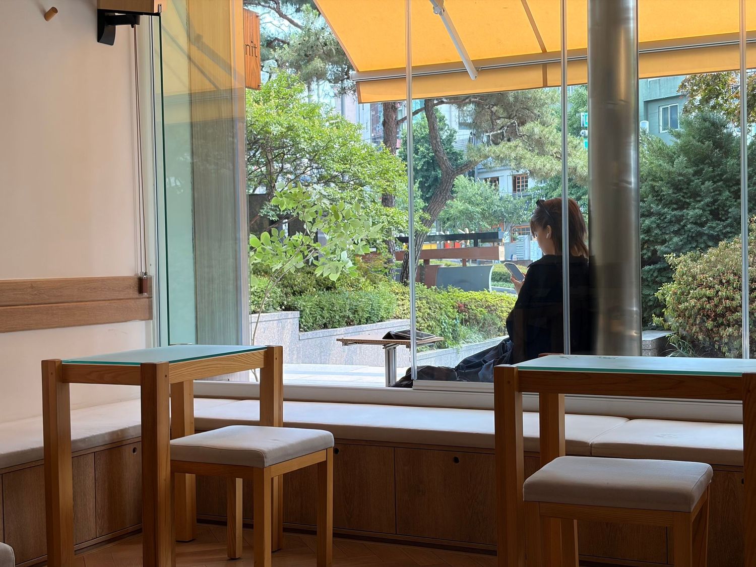Person sitting by large windows in a cozy wooden cafe with garden view and yellow awning.