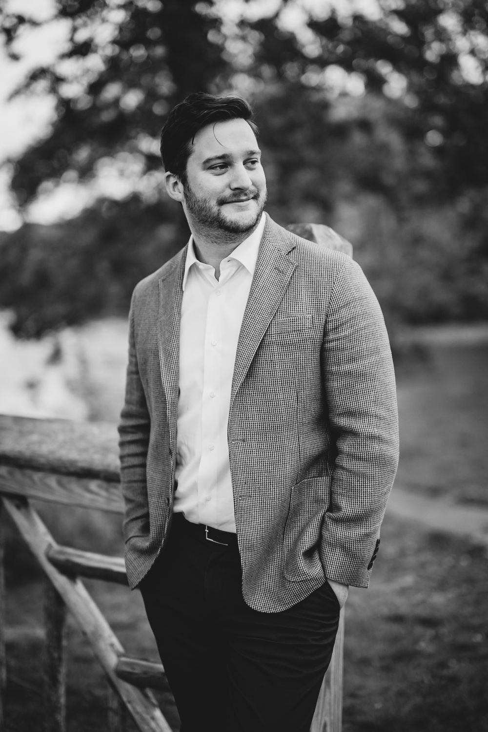 Black and white portrait of a smiling man in a blazer standing by a wooden fence outdoors.