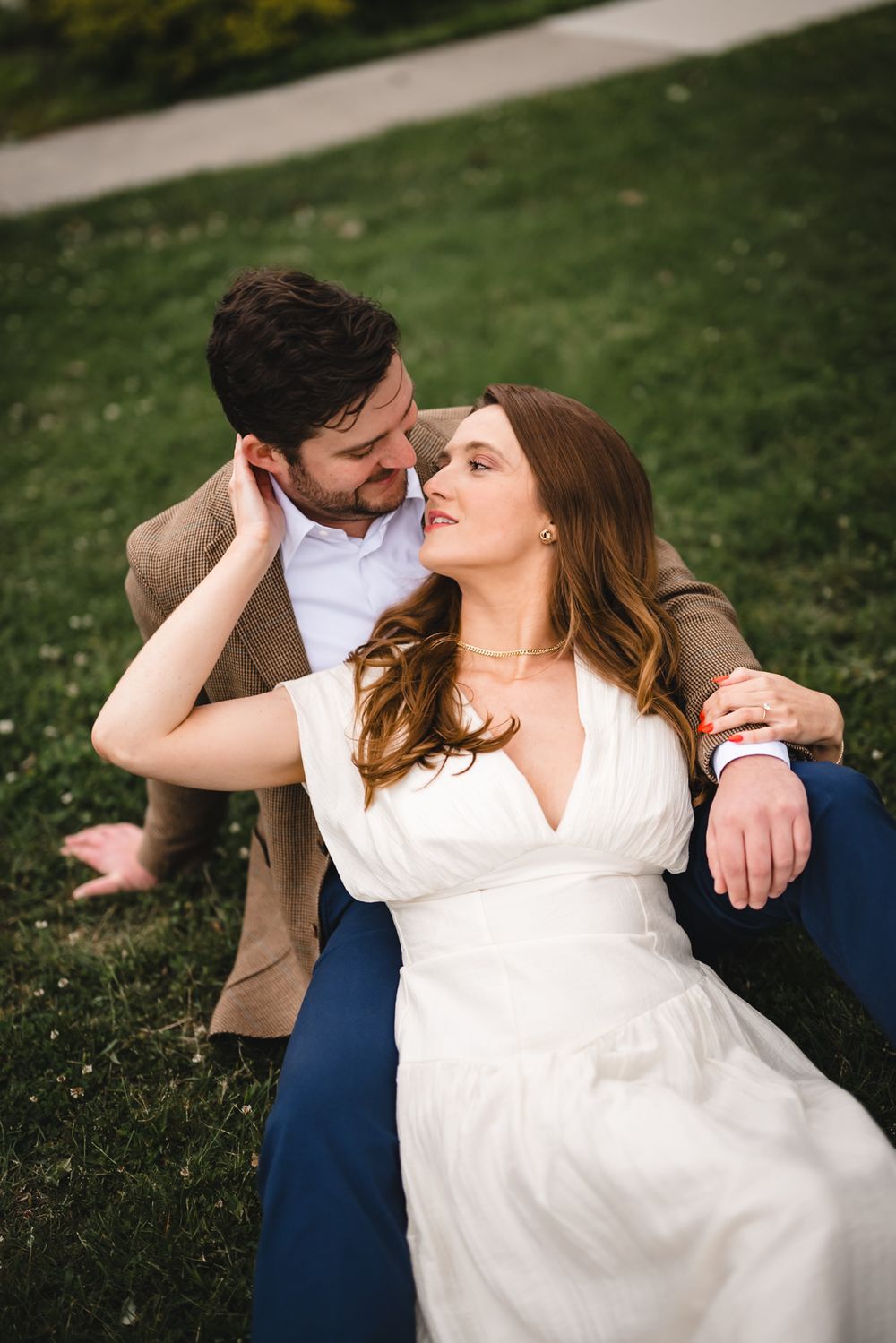 Couple sitting on green grass, woman in white dress leaning against man in brown blazer.