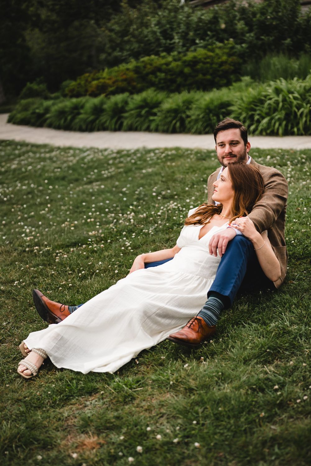 Couple sitting on green grass in a park, woman in white dress leaning against man in brown jacket.