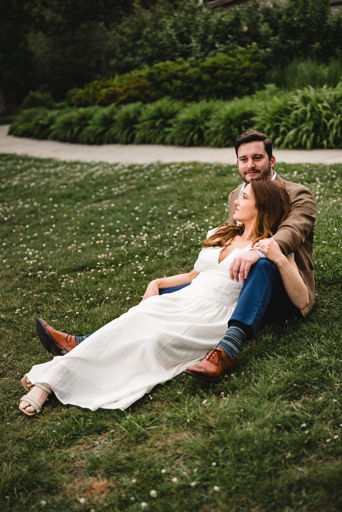 Couple sitting on green grass in a park, woman in white dress leaning against man in brown jacket.