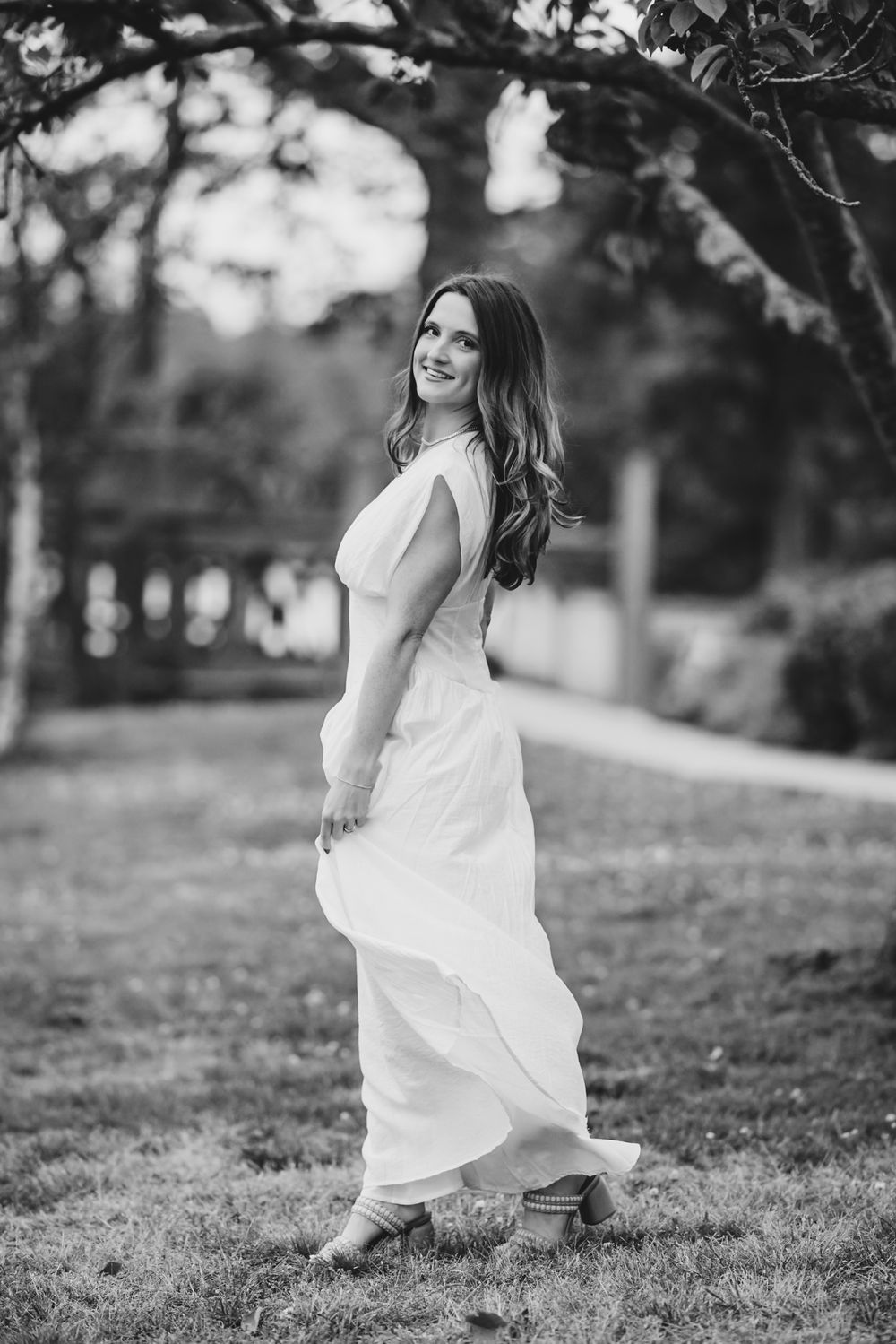 Black and white photo of a woman in a flowing white dress posing gracefully outdoors under a tree.