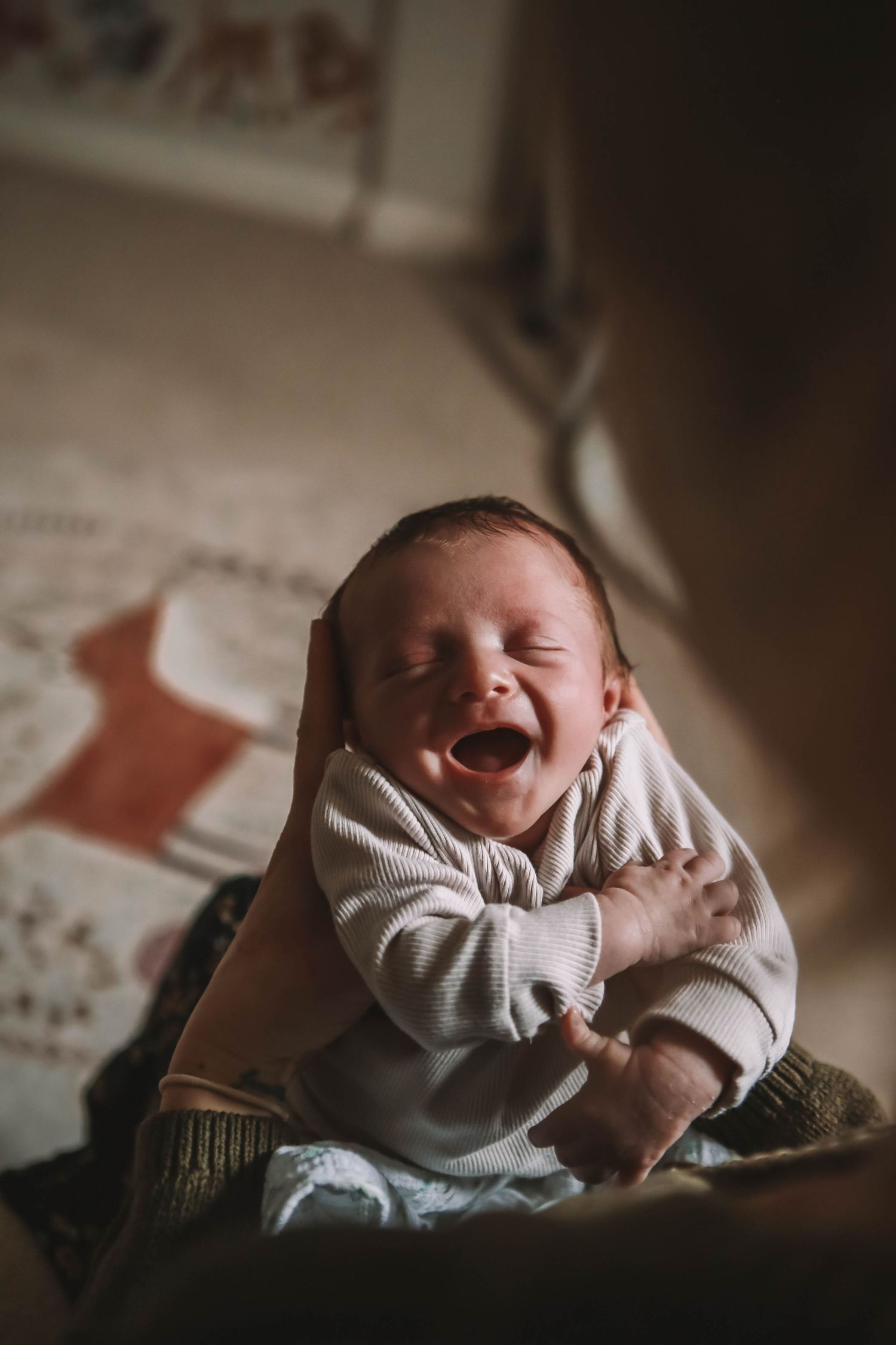 Newborn baby yawning while being held by a parent indoors, wearing a white striped onesie.