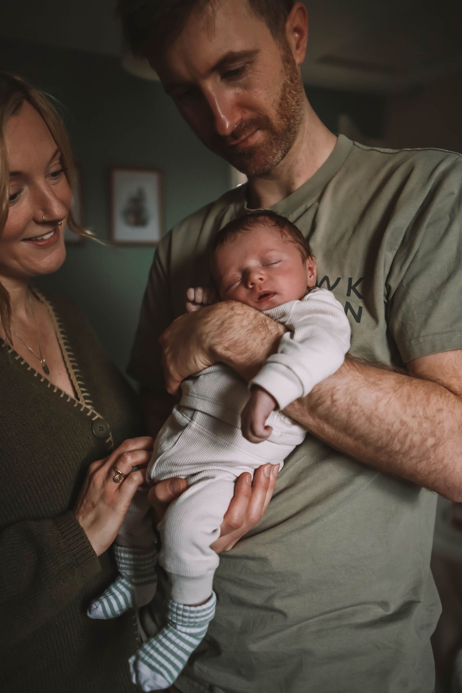 New parents smiling and cradling their sleeping newborn baby dressed in white at home.