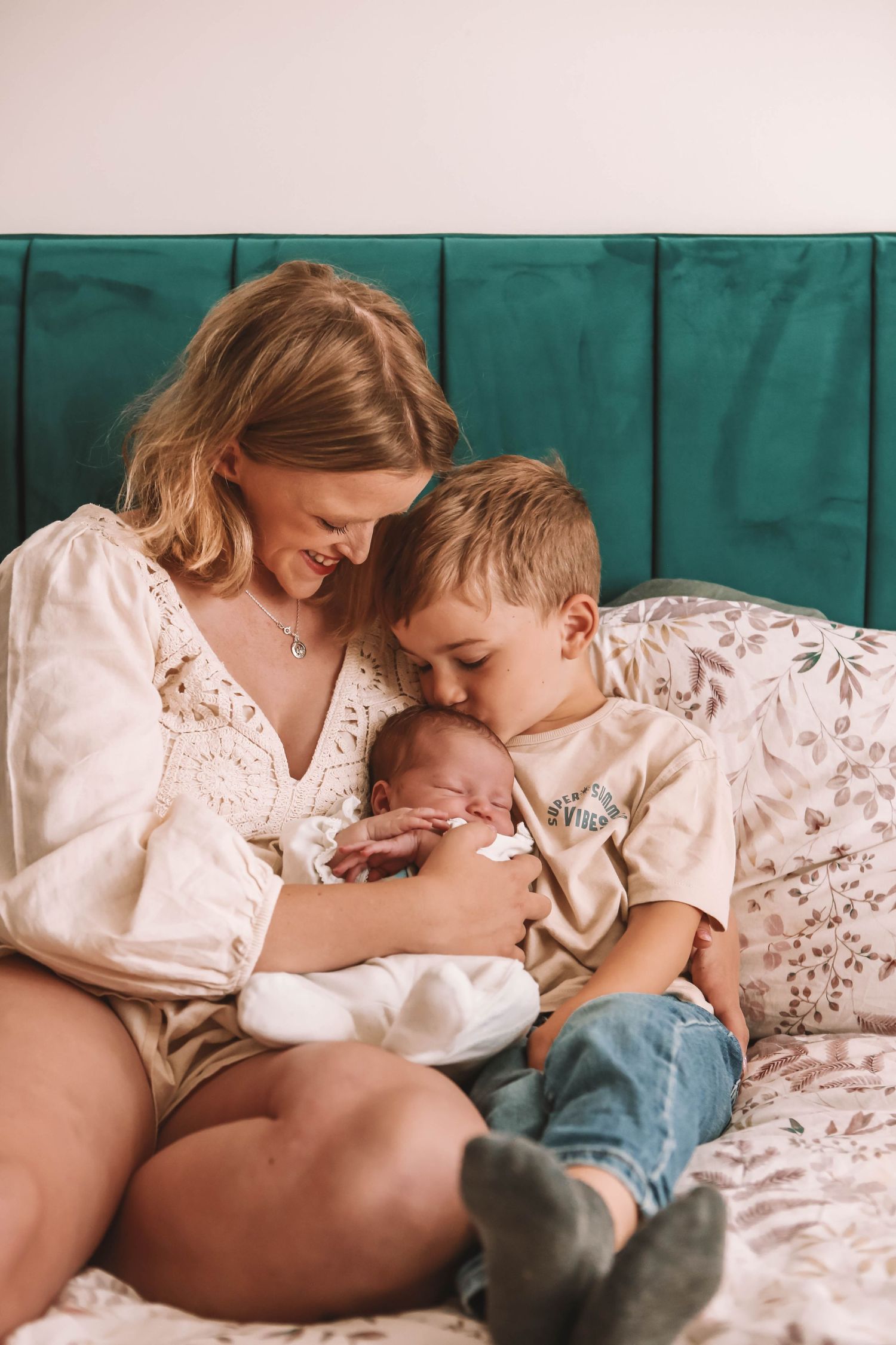 A smiling mother sits on a bed holding her newborn baby while her young son leans in to give the infant a gentle kiss.