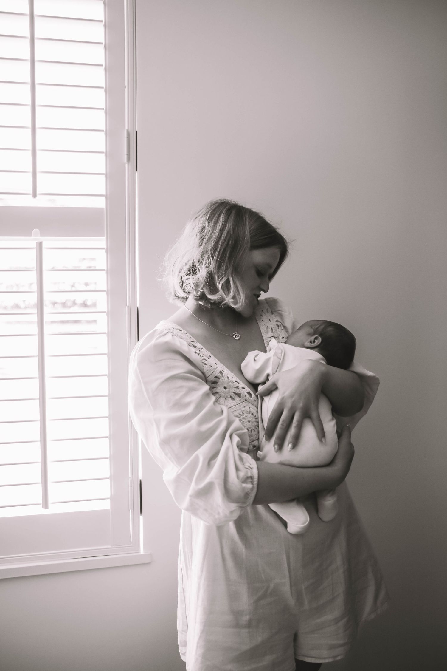 Black and white photo of a mother in white robe tenderly holding her newborn baby near a bright window.