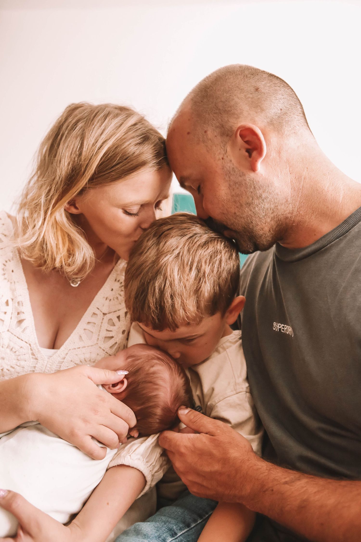 Family of four huddles together, parents touching foreheads while older child and newborn baby share a tender moment.