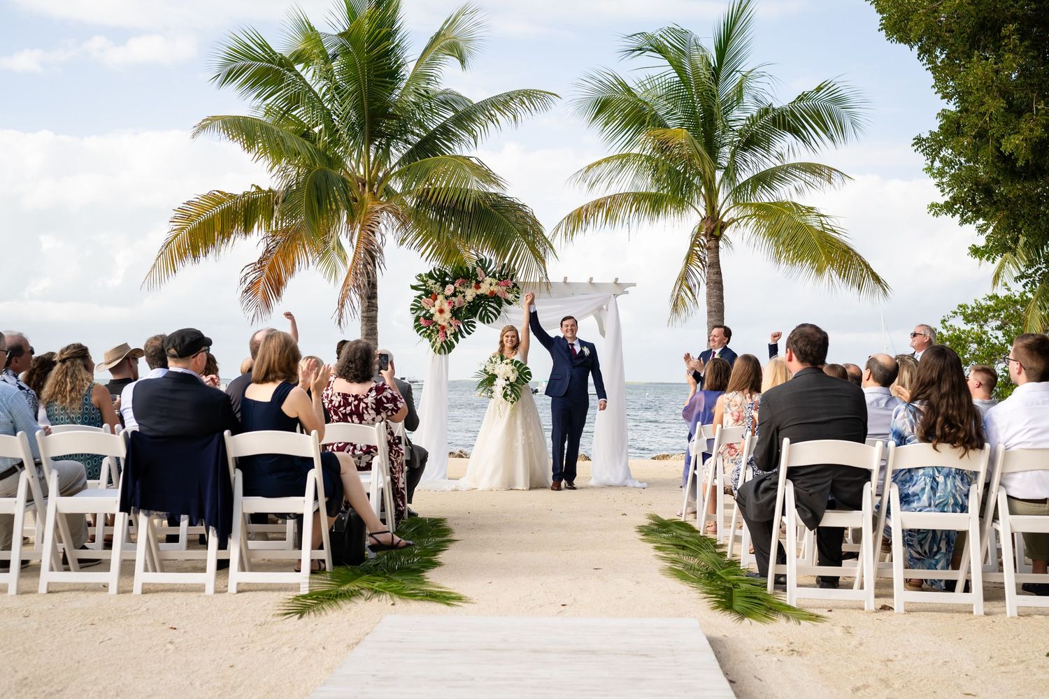 Newlyweds walk down beach aisle lined with white chairs and palm trees at an outdoor tropical wedding ceremony.