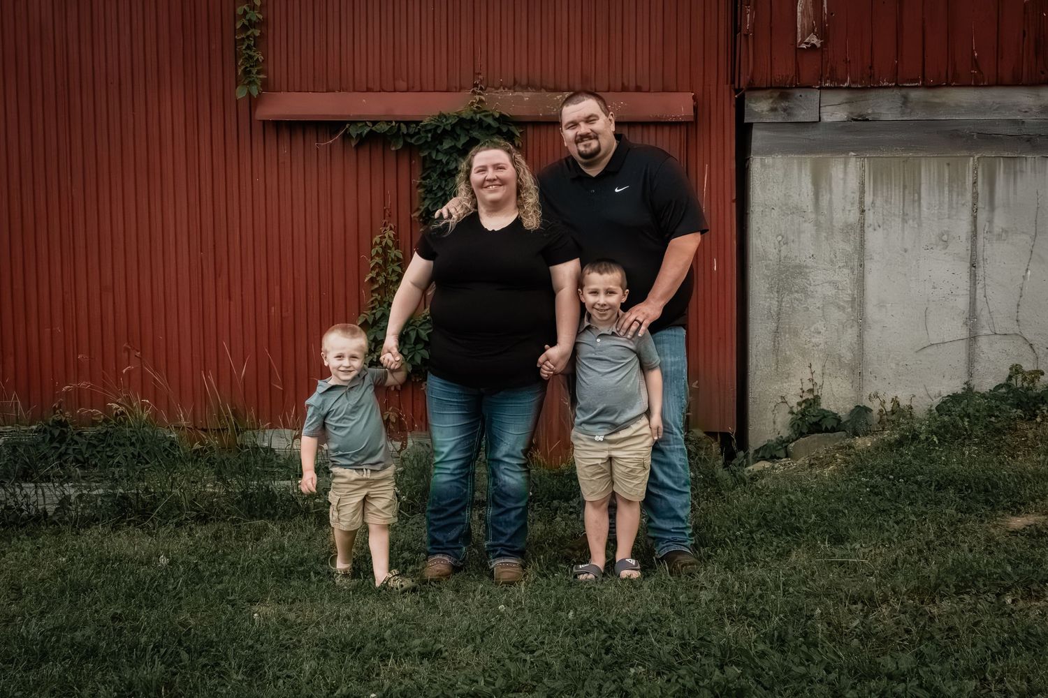 Family of four smiling together outdoors in front of a red barn, parents holding hands with two young boys.