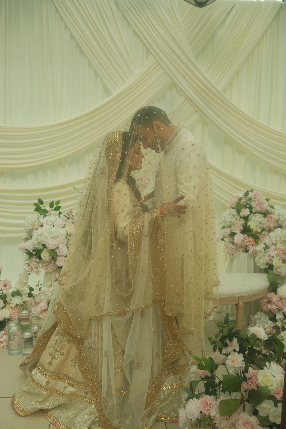 South Asian bride and groom in elegant gold and white traditional wedding attire sharing an intimate moment under draped fabric backdrop.