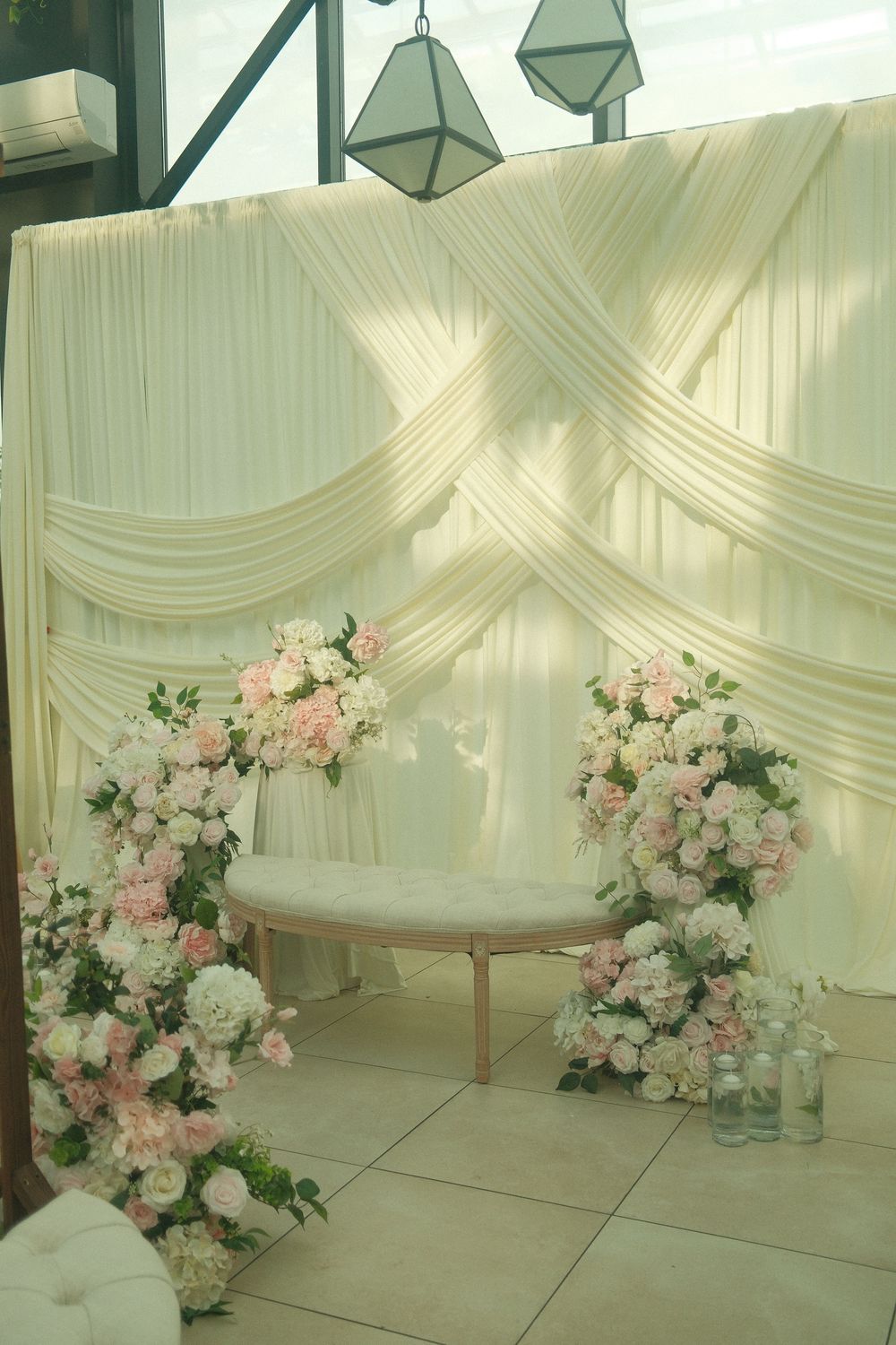 Elegant wedding backdrop with white draping, pink and white floral arrangements flanking a tufted bench on a tiled floor.