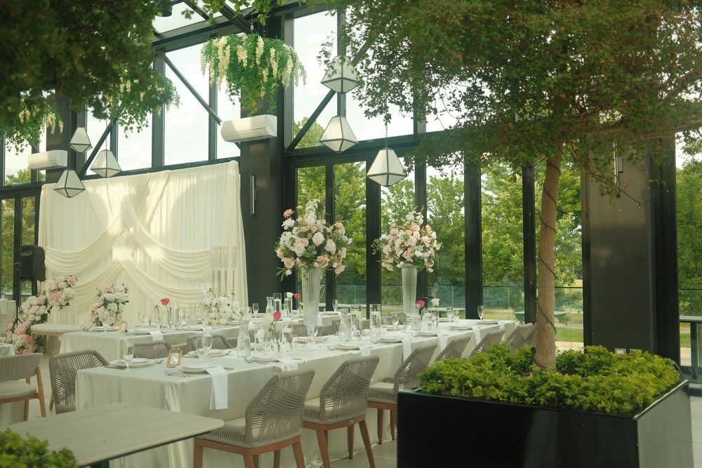 Elegant wedding reception in a glass venue with tall floral centerpieces, white linens, and lush greenery.