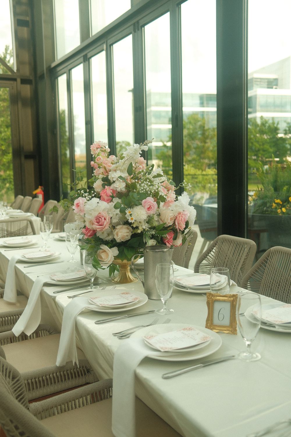 Elegant wedding reception table setting with pink and white floral centerpiece, white linens, and fine china near large windows.