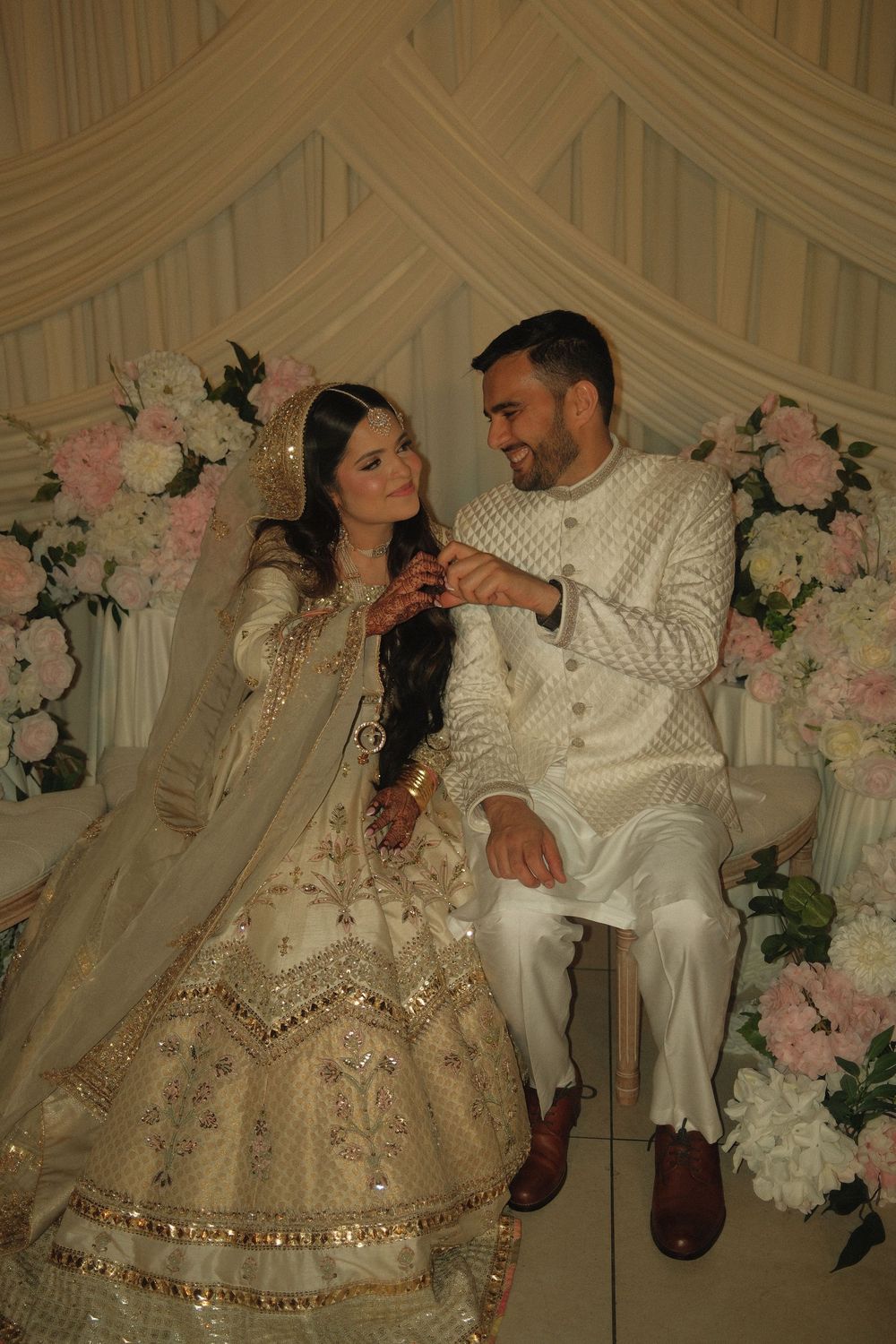 South Asian bride and groom in matching ivory outfits exchanging rings at their wedding ceremony with floral backdrop.