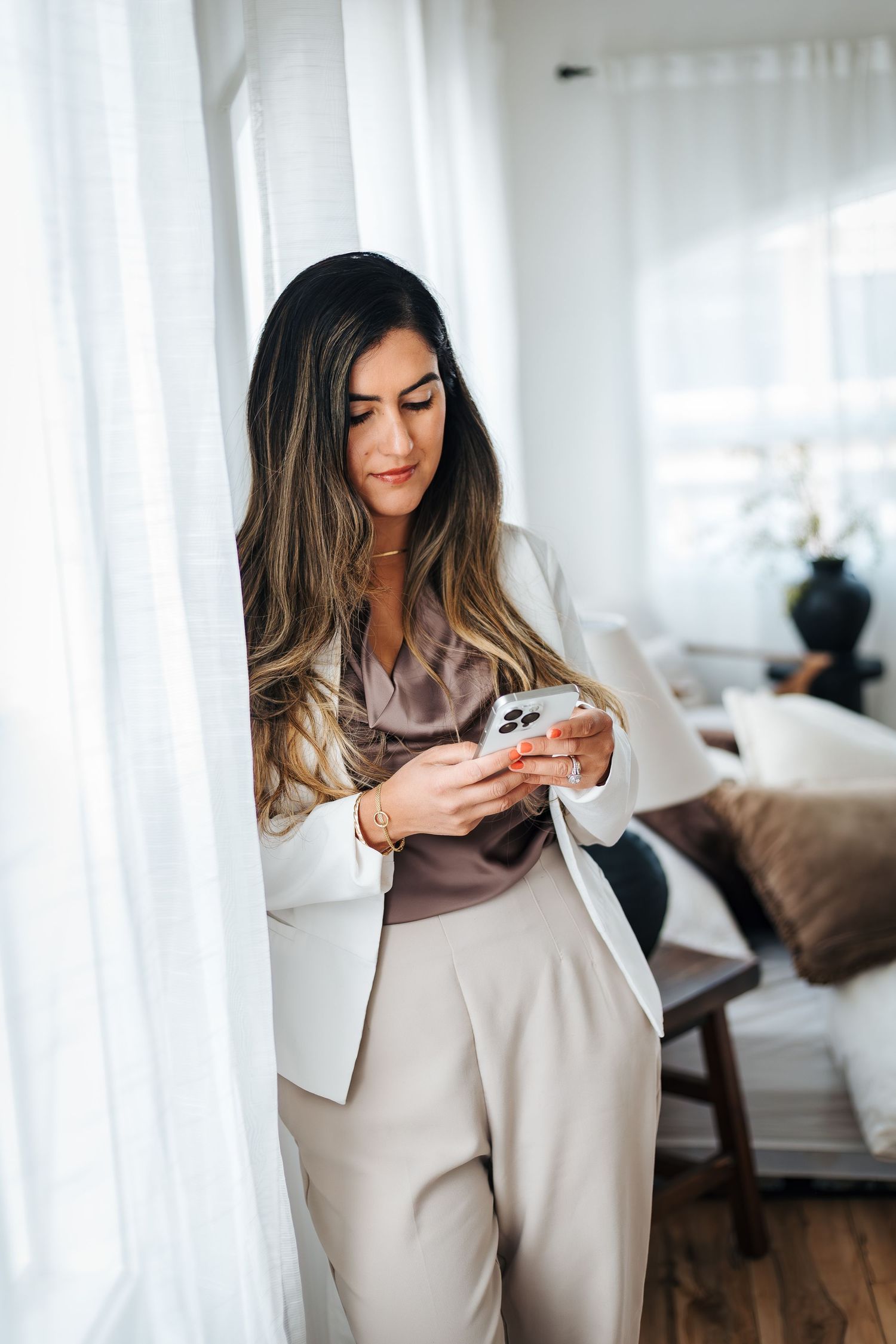Professional woman in blazer and beige pants using phone while leaning against window. Branding pictures and poses. Accountant headshots.