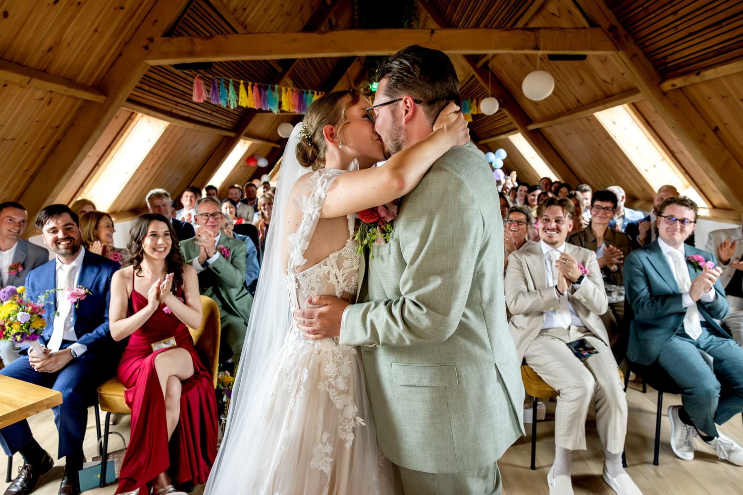 Bruidspaar deelt de eerste kus na het jawoord, met familie op de achtergrond tijdens de ceremonie bij Krelis in Uitgeest