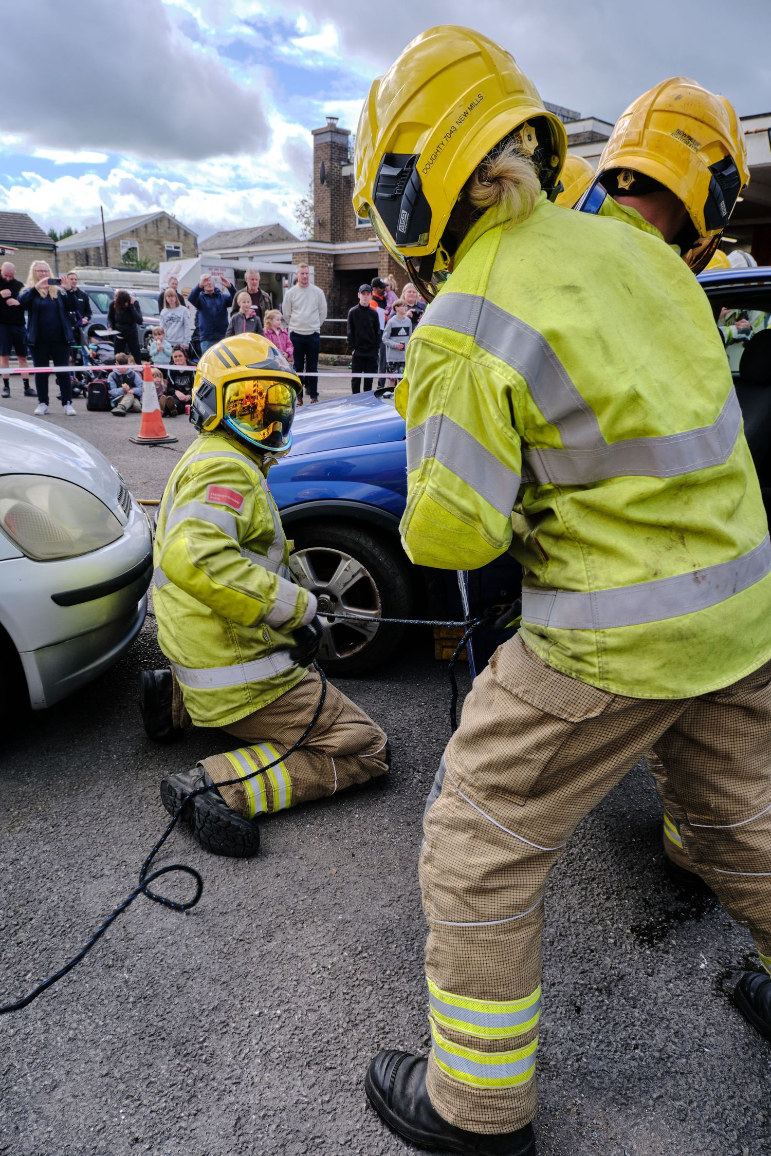Firefighters in yellow gear work on a crashed car during an emergency rescue demonstration watched by a crowd.