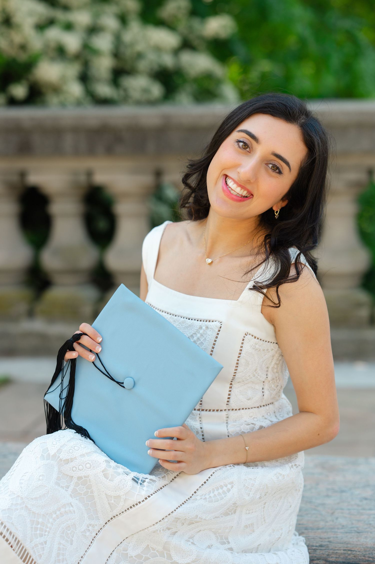 Smiling young woman in white lace dress holding a light blue graduation cap while sitting outdoors posing for Columbia graduation photos.