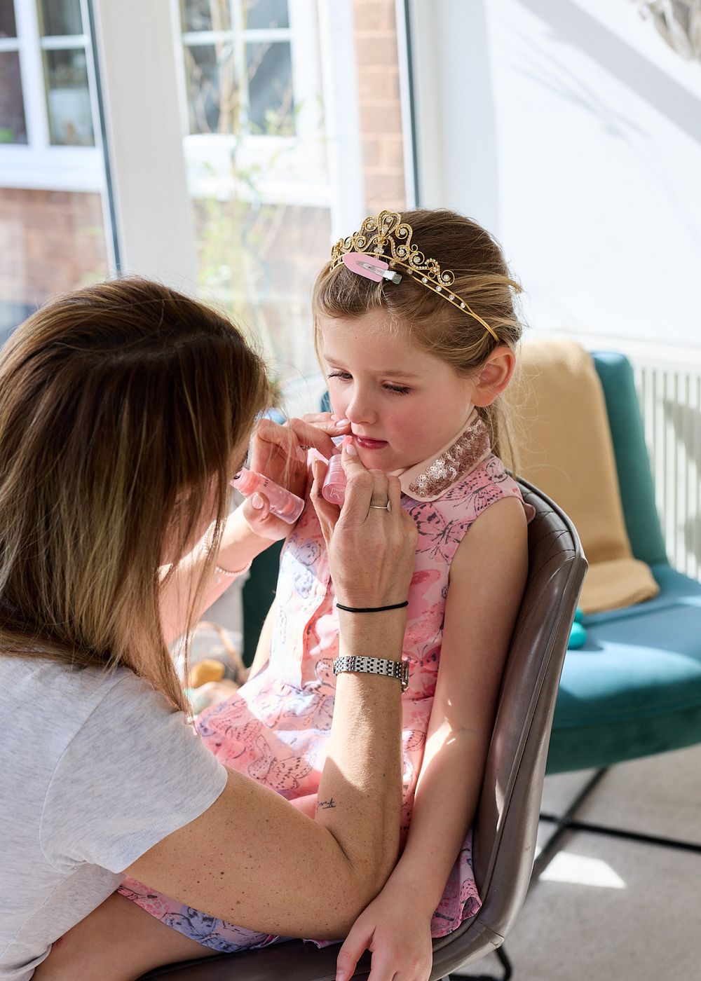 Woman applying lip makeup to a young girl wearing a gold tiara and pink dress at a party.