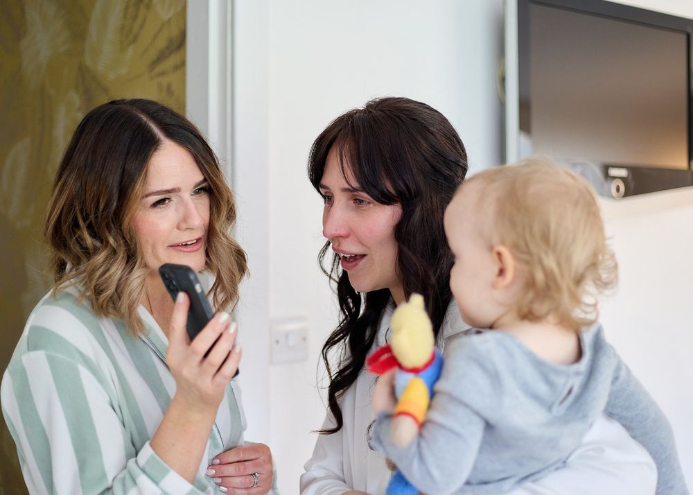 Two brunette women smiling at a phone while one holds a baby with a colorful toy indoors.