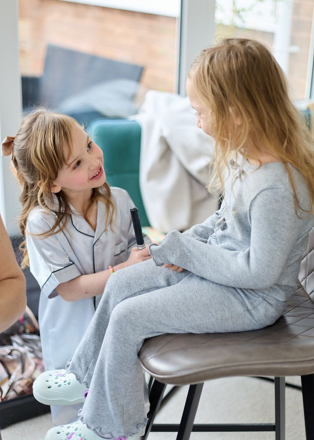 Two young girls in pajamas interact playfully, one holding a pen while sitting together in a bright living room.